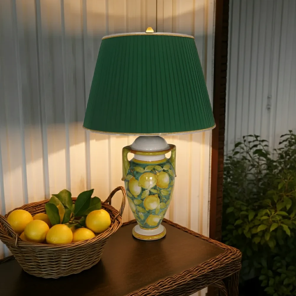 Paralume a tronco di cono in plissé color verde smeraldo Paralume a tronco di cono in tessuto plissé verde smeraldo, rifinito con bordi oro/bianco, montato su base in ceramica decorata con limoni.