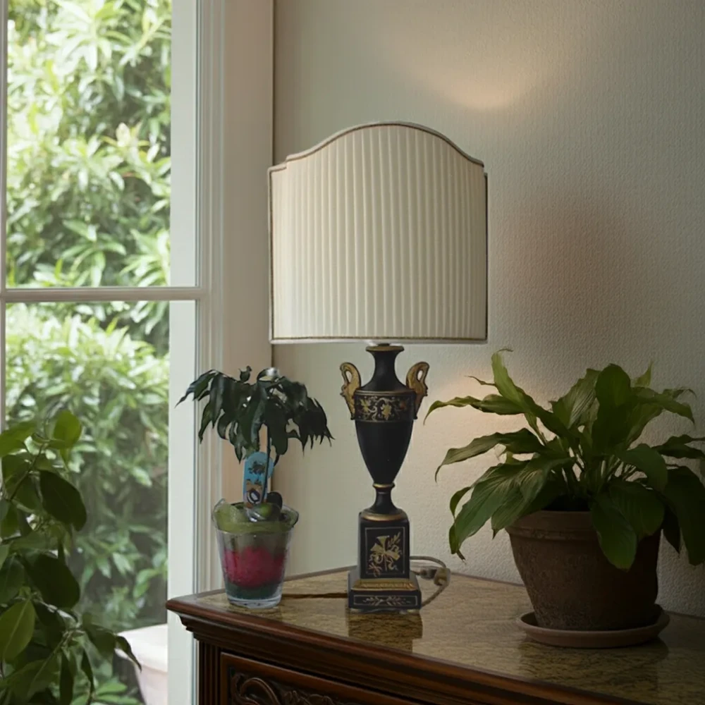 Paralume a Ventola in Stoffa a Pieghe Regolari per Lampada in Ceramica Decorata Paralume a ventola in stoffa color crema, cucita a pieghe regolari, montato su una lampada da tavolo con base in ceramica nera decorata con motivi dorati.