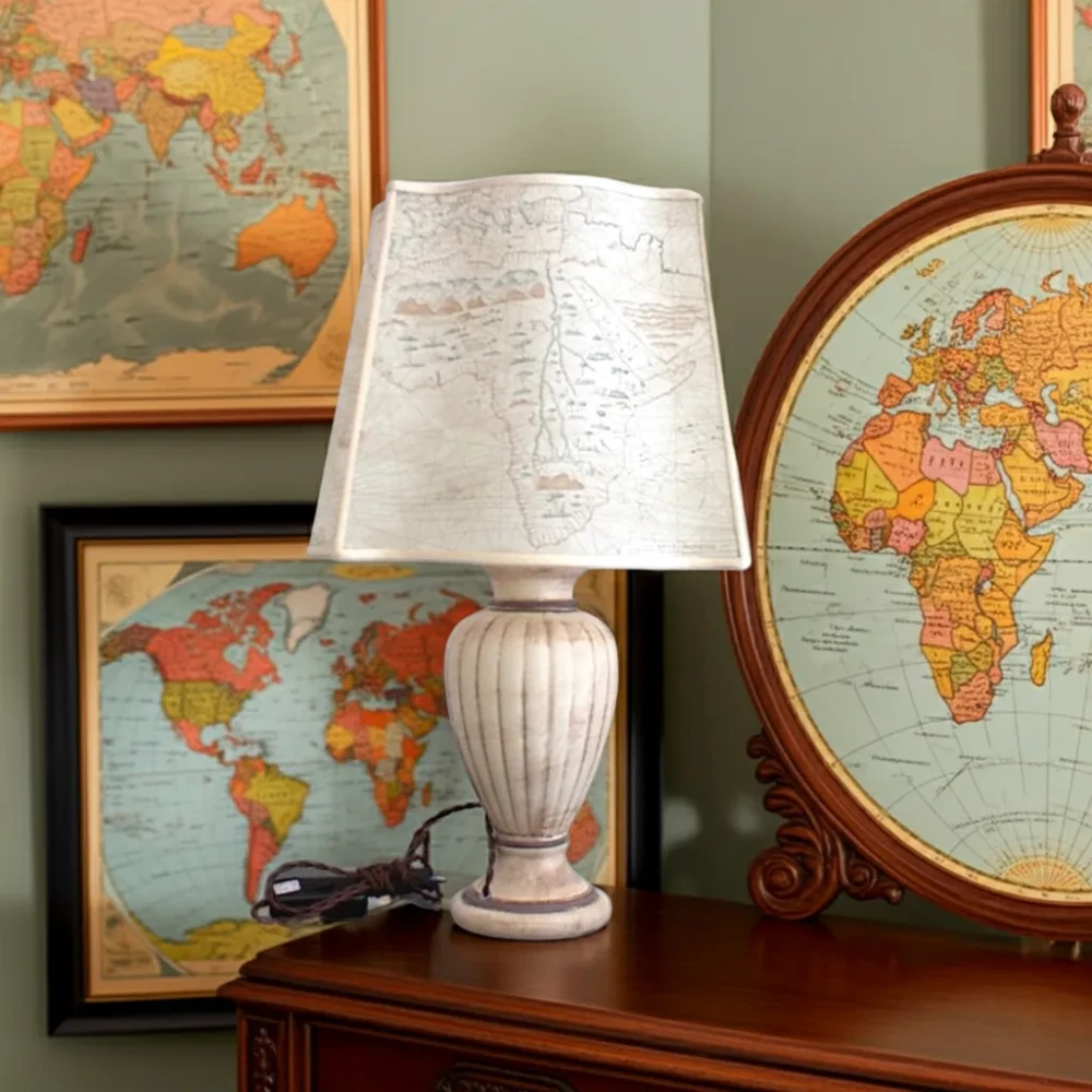 Paralume a Ventola con Lati Sagomati, Stampa Carta Geografica in Carta Pergamena Paralume con lati sagomati realizzato in carta pergamena con stampa di una mappa geografica antica, montato su una lampada da tavolo in stile classico con base color crema striata. L'oggetto è in un ambiente a tema viaggio con mappe incorniciate.