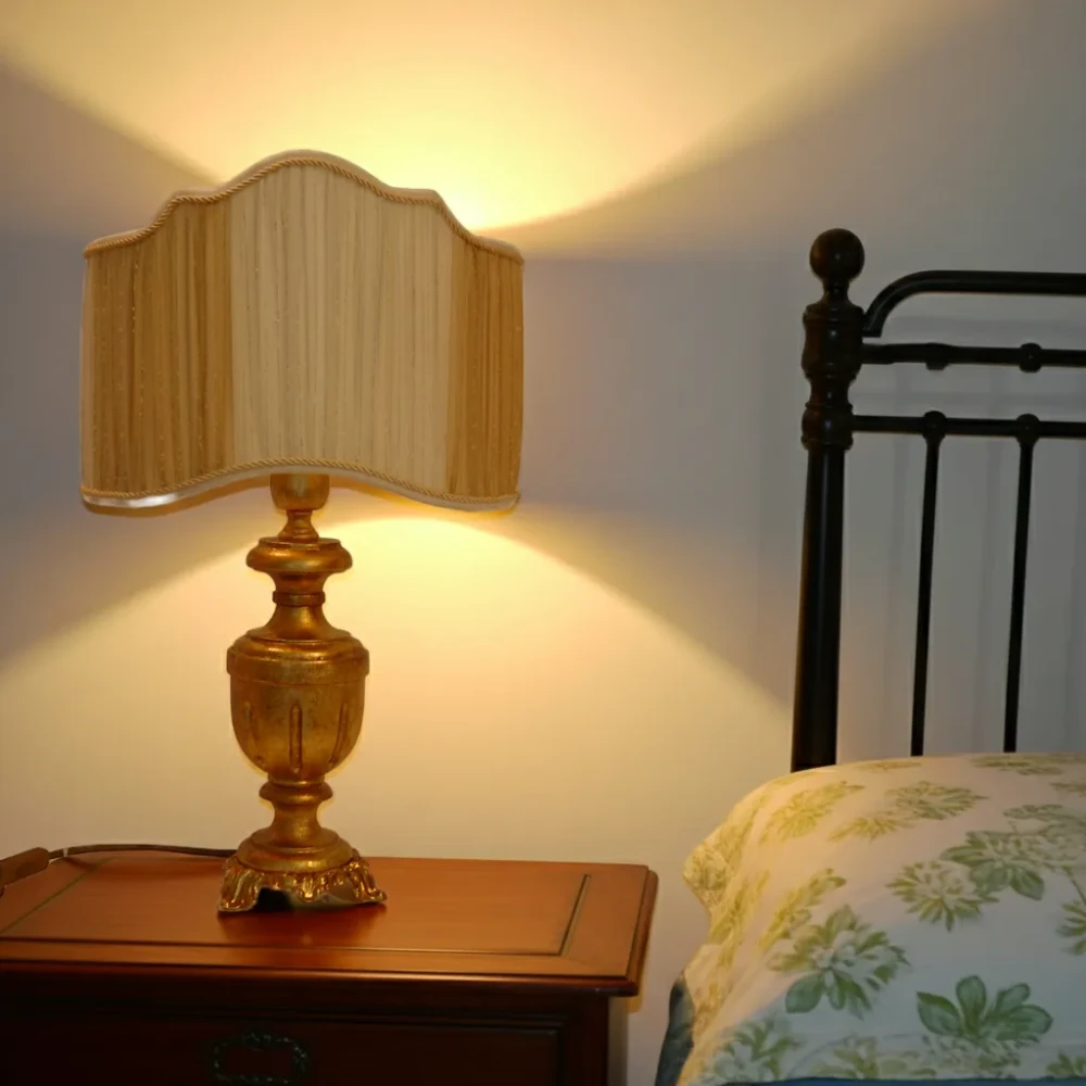 Paralume a Ventola Bicolore con Bordi Ondulati per Lampada da Tavolo Elegante paralume a ventola plissettato con bordi ondulati e finitura bicolore (oro/beige), montato su una lampada da tavolo con base dorata in stile classico su un comodino