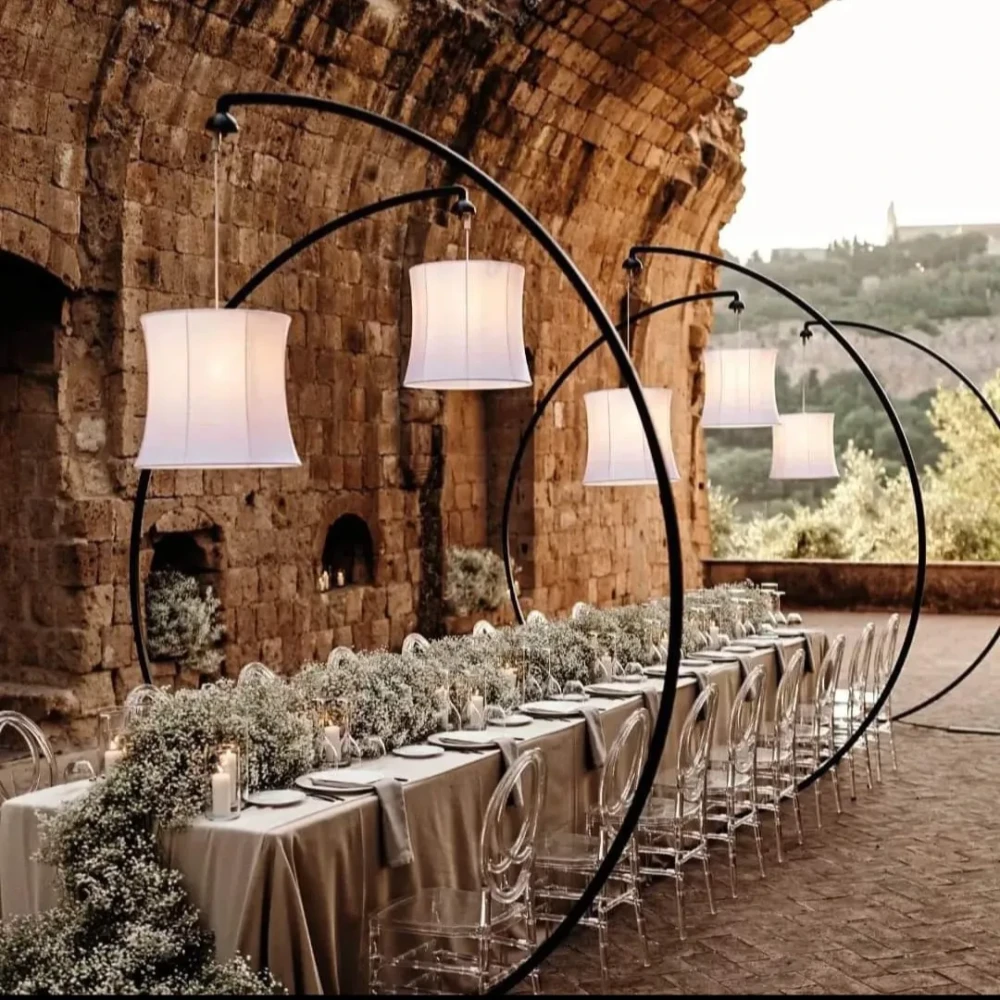Paralume Sospensione Cilindro Svasato Paralumi a sospensione di forma cilindro svasato, in tessuto bianco, montati su strutture ad arco per illuminare un lungo tavolo nuziale in un contesto rustico e monumentale.