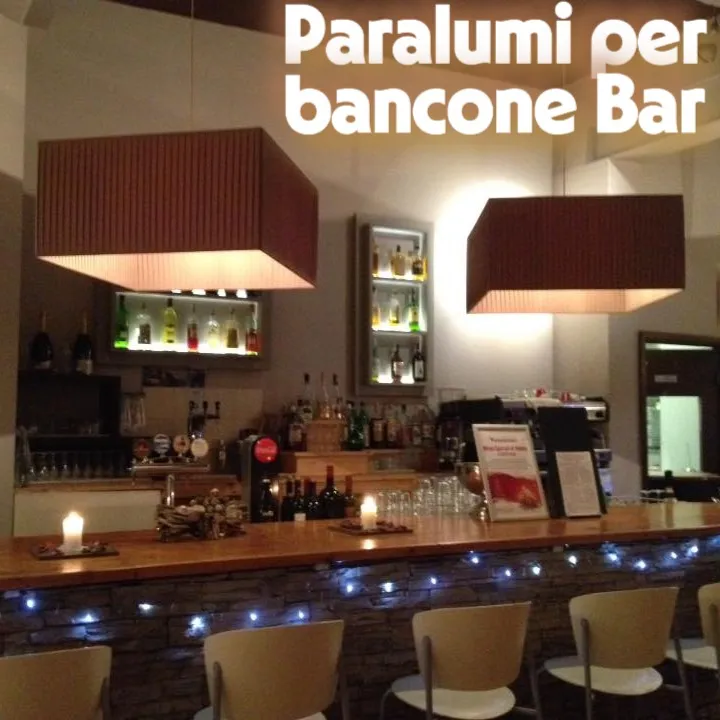 Paralumi Quadrati a Pieghe per Bancone Bar - Colore Tabacco Due grandi paralumi quadrati in tessuto a pieghe color tabacco, appesi sopra il bancone di un bar.