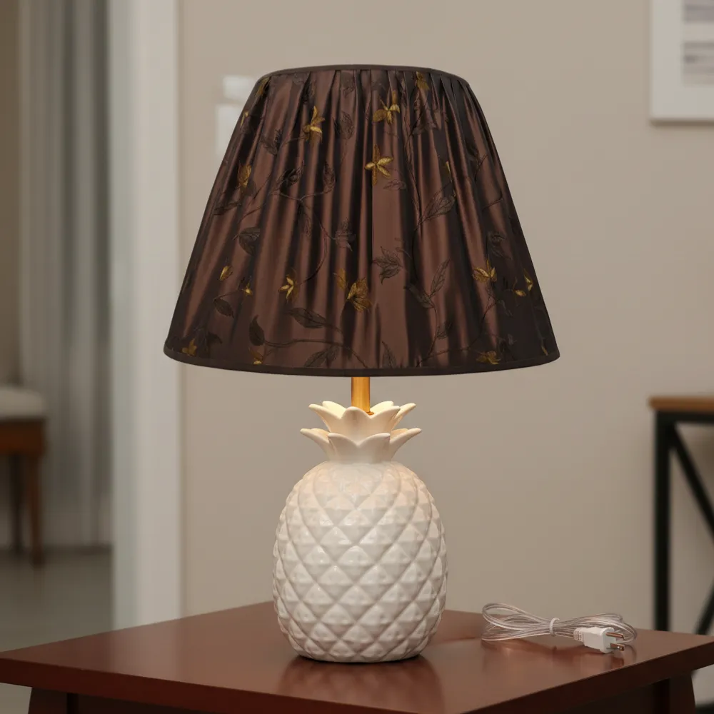 Paralume artigianale tronco conico in tessuto marrone ricamato su una moderna lampada da tavolo in ceramica bianca a forma di ananas.