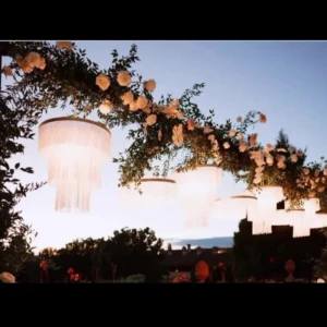Paralumi a sospensione a più strati, realizzati con frange lunghe e bianche, che creano un effetto chandelier e illuminano un evento all'aperto o un tavolo nuziale.